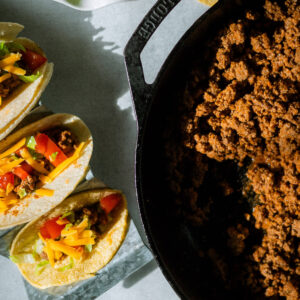 ground beef taco meat in a skillet, with tacos with ground beef, cheese, tomatoes and lettuce wrapped in a corn tortilla